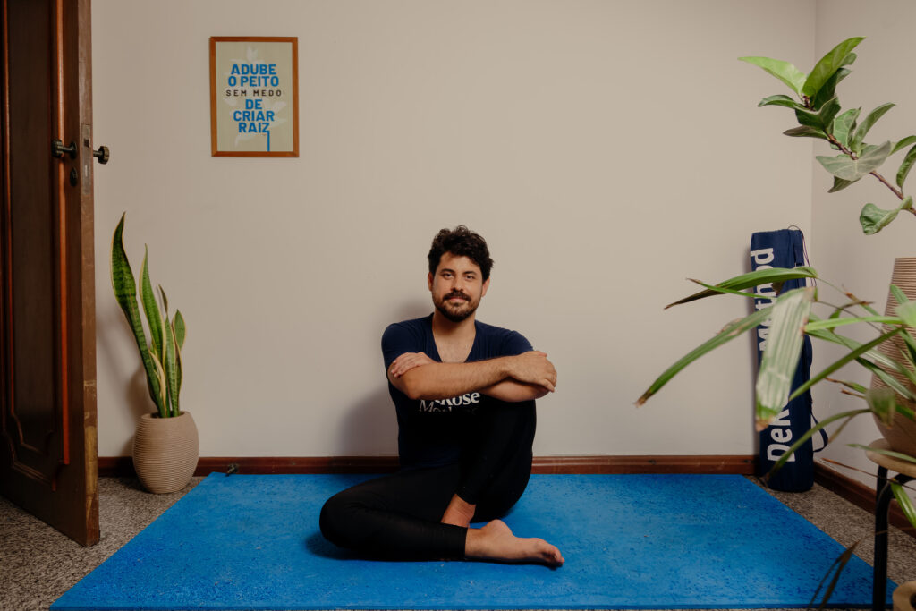 Homem sentado com as pernas cruzadas sobre uma placa de EVA azul com plantas à sua volta. Olhos abertos e leve sorriso no rosto. A imagem apresenta o contexto para a Meditação. 